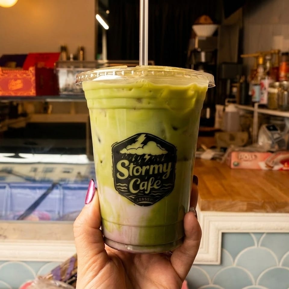 Hand holding an iced green and purple drink with a "Stormy Cafe" logo inside a cafe.