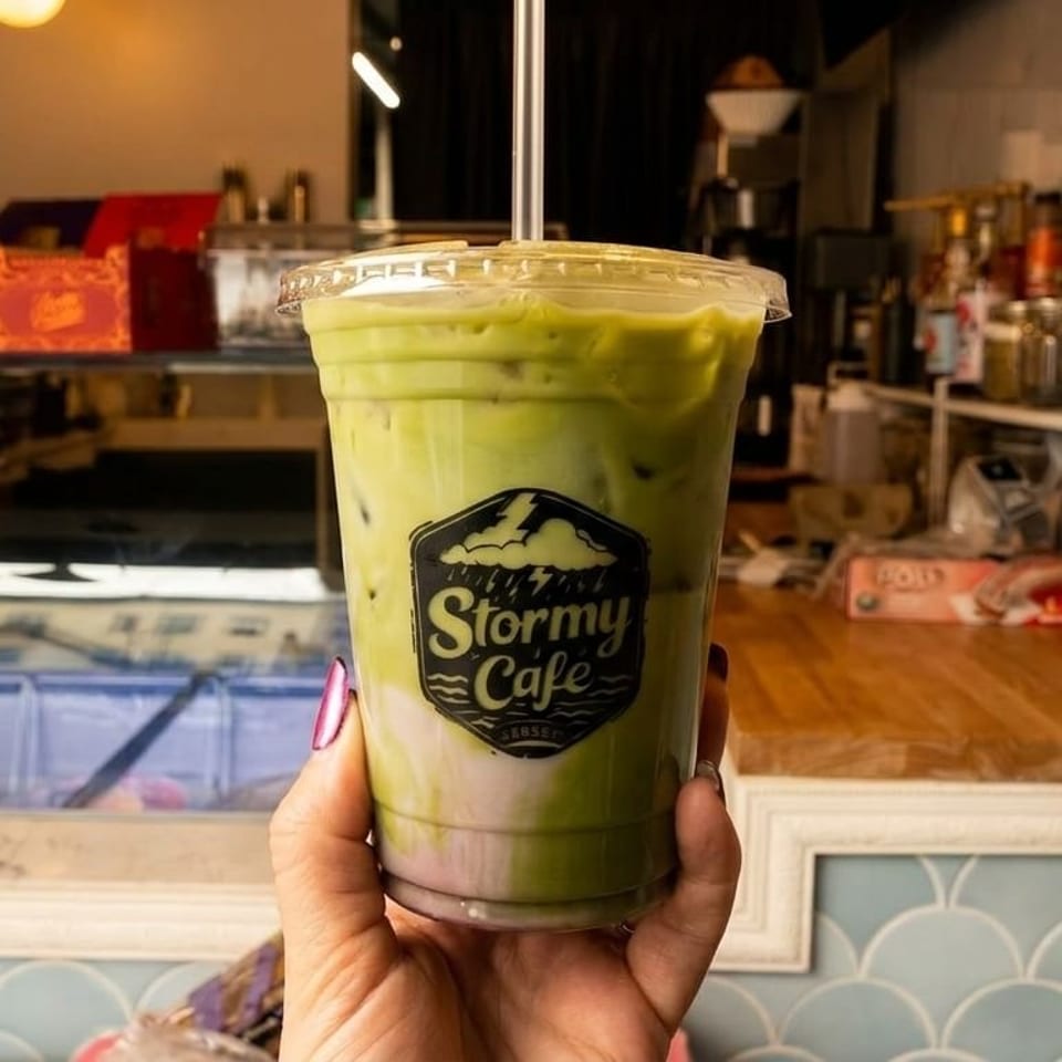 Hand holding an iced green and purple drink with the "Stormy Cafe" logo in a cafe setting.